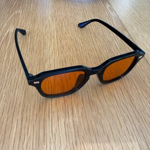 Helt nya solglasögon - Snygga solglasögon med svart, kantig båge och orangefärgade glas. Perfekt accessoar för att sticka ut i sommar. Bågarna har en modern, lite oversized look och passar de flesta ansikten.