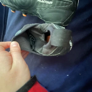 Nike Phantom GT fotbollsskor - Säljer ett par svarta Nike Phantom GT junior fotbollsskor med orange dobbar och vit Nike-logga. Skorna har en strumpliknande passform utan traditionell snörning och är designade för bra grepp på planen.