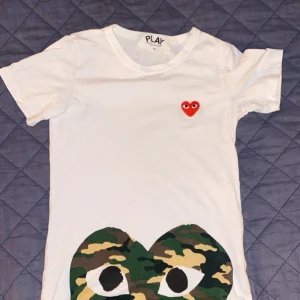 Vit t-shirt från Comme des Garçons Play med camo-hjärta - Vit t-shirt från Comme des Garçons Play med det klassiska röda hjärtat på bröstet och ett stort camo-mönstrat hjärta med ögon framtill. Rund halsringning och korta ärmar. Perfekt statement-plagg för dig som gillar streetwear. Det står S men sitter mer som XS