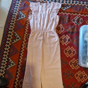 Ljusrosa volang bodysuit från Lindex - Supersöt ljusrosa bodysuit från Lindex med volangdetaljer på axlarna och resår i midjan. Klänningen har rund halsringning och dragkedja bak. Perfekt för somriga dagar eller speciella tillfällen.