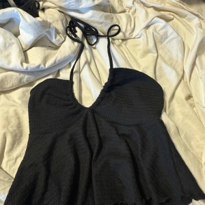 Svart croppad linnetopp med knyt - Säljer en svart croppad linnetopp med smockad struktur och volangkant. Toppen har smala axelband som kan korsas och knytas i nacken för en snygg detalj. Materialet är mjuk polyester med lite stretch. Perfekt för dig som gillar trendiga och bekväma toppar.