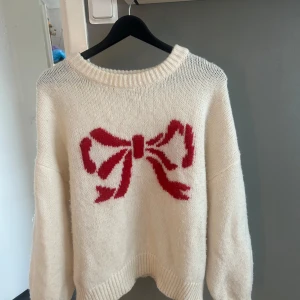 Vit stickad tröja med röd rosett - Superfin vit stickad tröja med en stor röd rosett på bröstet. Tröjan har rund hals och långa ärmar med ribbade muddar. Perfekt för dig som gillar gulliga detaljer och vill ha en mysig look. Passar extra bra under höst och vinter. Storlek large men passar s-m bäst