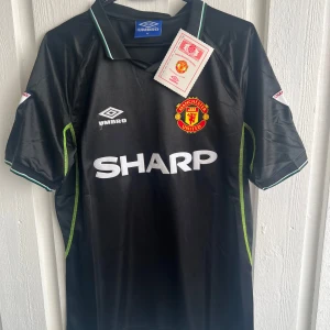 Manchester United Scholes 18 Umbro tröja - Säljer en svart Manchester United fotbollströja från Umbro med Scholes 18 på ryggen. Tröjan har korta ärmar, vita tryck och gröna detaljer längs sidorna och ärmslut. Klassisk krage och klubbmärke på bröstet. Materialet är glansigt polyester.