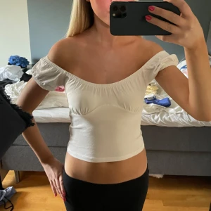 Vit croppad offshoulder topp - Supersöt vit croppad topp med offshoulder och puffiga korta ärmar. Tunn och mjuk i bomull, med snygg rynkning vid halsen och under bysten som ger en feminin vibe. Perfekt till svarta jeans eller kjol för en chill och trendig look.