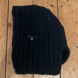 Svart polotröja från GANT (tjej) - Svart polotröja från GANT med kabelstickat mönster och broderad logga på bröstet. Tröjan är tillverkad i stretchig bomull och har lång ärm samt hög polokrage. Perfekt för dig som gillar klassisk och stilren look.   STORLEK- S