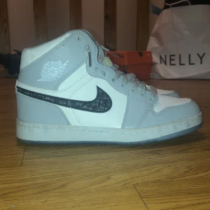 Nike Air Jordan 1 x Dior sneakers - Säljer ett par Nike Air Jordan 1 x Dior sneakers i grått och vitt med svart Dior-mönstrad swoosh. Skorna har högt skaft, snörning och en transparent sula med Dior-logga undertill. Perfekt för dig som vill sticka ut med en riktigt ikonisk collab.