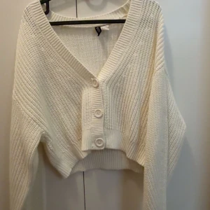 Vit croppad kofta från hm - Säljer en vit croppad kofta från hm med grovstickad struktur och bred ribbad kant. Koftan har djup v-ringning, tre stora knappar framtill och långa ärmar. Perfekt för en mysig och varm och samtidig snygg stil.