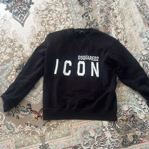 Svart sweatshirt Dsquared2 ICON - Svart sweatshirt från Dsquared2 i storlek M med stor vit 'ICON'-print på bröstet och Dsquared2-logga. Tröjan har rund hals, långa ärmar och ribbade muddar. Perfekt för dig som gillar streetwear och vill sticka ut med en snygg statement-tröja. Top kvalite