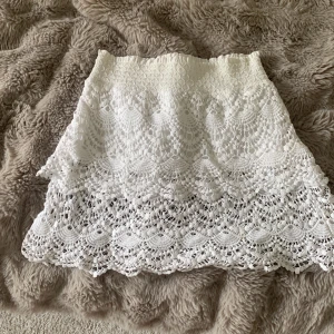 Vit virkad kortkjol med volang - Superfin vit kortkjol med virkad spets och volangdetaljer. Kjolen har elastisk midja och ett snyggt mönster som ger en romantisk vibe. Perfekt för sommarens alla tillfällen och lätt att matcha med både sneakers och sandaler.