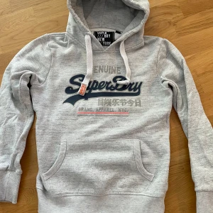Grå hoodie från Superdry XS - Säljer en grå hoodie från Superdry i storlek XS. Tröjan har huva med snörning, stor Superdry-logga på bröstet och en praktisk magficka. Mjuk insida och ribbade muddar vid ärmar och nederkant. Perfekt för chill dagar och streetstyle. Kontakta vid fundering och priset kan diskuteras🙌