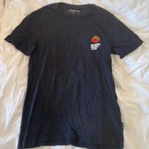 Svart HRTBRKR Social Club t-shirt - Svart t-shirt från YOURTURN med HRTBRKR Social Club-tryck. Framsidan har ett litet hjärta med eld och text, medan ryggen har ett stort motiv med rött hjärta och pilar. Skön passform och tillverkad i mjuk bomull. Perfekt för dig som gillar coola prints och streetstyle.