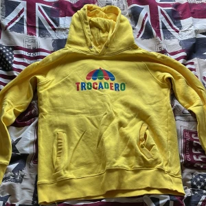 Gul Trocadero hoodie med färgglad logga - Säljer en gul hoodie från Trocadero med en stor, färgglad logga och paraply på bröstet. Tröjan har huva med snörning, ribbade muddar och två fickor framtill. Perfekt för dig som gillar starka färger och vill sticka ut lite extra.