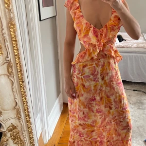 Zara Blommig långklänning med volang - Säljer en färgstark långklänning med blommigt mönster i rosa, orange och gult. Klänningen har djup v-ringning, volangdetaljer och öppen rygg med knytband. Tillverkad i lätt linne, perfekt för varma dagar. Snygg och unik design som sticker ut.