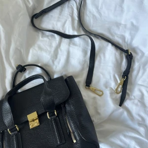 Svart väska från 3.1 Phillip Lim - Snygg svart väska i skinn från 3.1 Phillip Lim med guldiga metalldetaljer och dragkedjor framtill. Väskan har både handtag och avtagbar axelrem, samt ett rymligt fack med innerficka. Perfekt för dig som gillar stilrena och lyxiga accessoarer