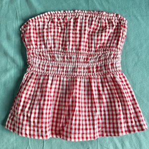 Rutig röd tubtopp från Stradivarius - Säljer en söt rutig tubtopp från Stradivarius i storlek M. Toppen är vit och röd med smockad midja och volang nedtill. Den är axelbandslös och har en bandeauformad siluett, perfekt för varma dagar. Materialet är mjuk bomull som känns skönt mot huden.