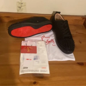 Christian Louboutin svarta sneakers med nitar - Säljer ett par svarta sneakers från Christian Louboutin med ikonisk röd sula och coola svarta nitar på tån. Skorna har snörning och är tillverkade i mocka för en lyxig vibe. Perfekt för dig som vill sticka ut med en edgy och exklusiv look.