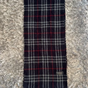 Rutig mörkblå halsduk från Burberrys - Snygg halsduk från Burberrys i klassiskt rutigt mönster med mörkblå, grå och röda toner. Gjord av 100% lammull, mjuk och varm med fransar i ändarna. Perfekt för att lyfta din stil under kyliga dagar.