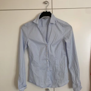Ljusblå skjorta från H&M, strl 36 - Snygg ljusblå skjorta från H&M i storlek 36. Klassisk krage, långärmad med knappar vid manschetterna och framtill. Skjortan är i mjuk bomull och har en tight passform som sitter bekvämt. Perfekt för dig som gillar stilrena och enkla plagg.