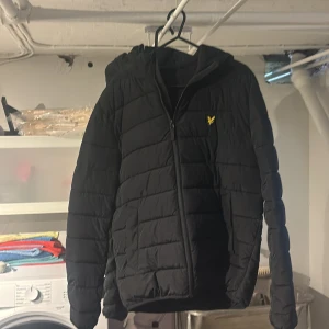 Svart pufferjacka från Lyle & Scott - Svart pufferjacka från Lyle & Scott med huva och gul logga på bröstet. Jackan har quiltad design, dragkedja framtill och långa ärmar. Perfekt för kyliga dagar och har en clean, stilren look som funkar till streetwear. Materialet är polyester.