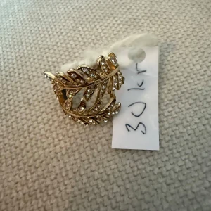 Guldig ring med glittriga stenar - Säljer en bred guldig ring med ett snyggt blad-inspirerat mönster. Ringen är dekorerad med små glittrande stenar som ger extra bling. Perfekt statement piece för dig som vill sticka ut med accessoarerna.