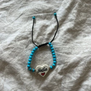 Blått pärlarmband med hjärta - Snyggt armband med blå pärlor och ett vitt hjärthänge där det står 'Julia' och en liten röd hjärtsymbol. Armbandet har svart justerbart snöre och silverfärgade detaljer vid hänget. Perfekt accessoar för att ge din stil en personlig touch.