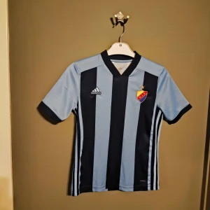 Djurgården fotbollströja Adidas  - Snygg Djurgården fotbollströja från Adidas i storlek barn M. Tröjan har breda marinblå och ljusblå ränder, korta ärmar och V-ringad krage. Tillverkad i lätt och ventilerande Climalite-material, perfekt för match eller träning. DIF-emblem på bröstet och Adidas-logga. Tröjan är 2016/17 säsongens hemma tröja