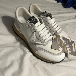 Valentino Garavani Rockrunner sneakers - Snygga Valentino Garavani Rockrunner sneakers i vitt och beige med coola nitar på hälen. Skorna har en mix av skinn och mocka, platt sula i gummi och klassisk snörning. Perfekt för dig som vill ha en clean och lyxig look med street vibe.