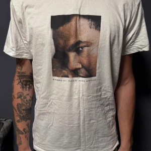 Vit t-shirt med tryck från Dedicated ”Muhammad Ali” - Säljer en vit t-shirt från Dedicated i storlek L, tillverkad av 100% ekologisk och Fairtrade-certifierad bomull. T-shirten har ett stort grafiskt tryck på bröstet och klassisk rund hals. Passformen är normal och materialet känns mjukt mot huden. Modellen på bilden är 190 cm lång och bär storlek L. Finns 2 stycken i lager.