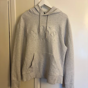 Grå hoodie från Hugo Boss - Säljer en stilren ljusgrå hoodie från Hugo Boss med broderad logga på bröstet. Tröjan har huva med snörning, stor magficka och långa ärmar. Perfekt för chill dagar och enkel att matcha med jeans eller joggers.