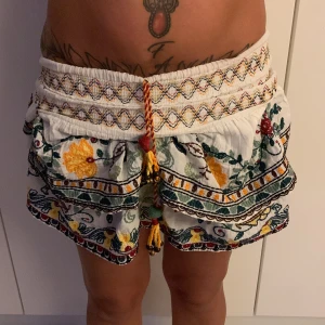 Broderade vita shorts med snörning - Unika vita shorts med färgglad broderad design i gult, grönt och rött. Högmidjade med bred resår och dekorativ snörning framtill. Volangdetaljer och mönstrade kanter ger en bohemisk vibe. Perfekta för sommarens alla äventyr! 