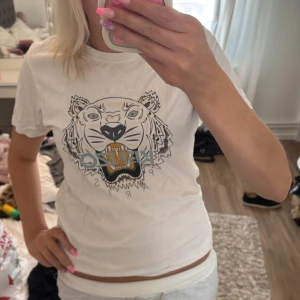 Vit Kenzo t-shirt med tigertryck - Säljer en vit t-shirt från Kenzo i storlek M med ikoniskt tigertryck och Kenzo Paris-logga på bröstet. T-shirten har rund hals, korta ärmar och är gjord i mjuk bomull. Perfekt statement-plagg med snyggt grafiskt motiv i svart, grått och gult.