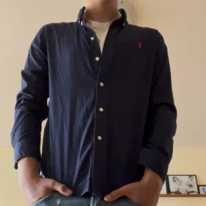Mörkblå skjorta från Ralph Lauren - Snygg mörkblå skjorta från Ralph Lauren med klassisk krage och vita knappar framtill. Liten röd logga broderad på bröstet. Skjortan är långärmad och har en normal passform, perfekt till jeans. Materialet är mjuk bomull som känns skönt mot huden. Storlek S, fraktar inom 48 h. Pris ej hugget i sten vid snabb affär!