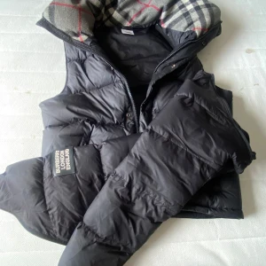 Svart väst/jacka från Burberry - Säljer en svart dunväst från Burberry med klassiskt rutigt foder i grått, svart och rött. Västen har dragkedja, knappar och två fickor med dragkedja framtill. Stor Burberry-patch på ena ärmen. Perfekt för kalla dagar och har en cool, puffig look.  Du kan även gör om jackan till en väst perfekt under hösten eller vår storleken är M men sitter med som S 