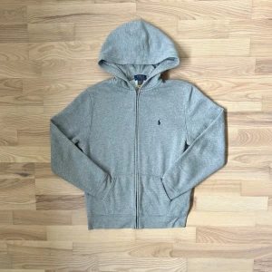 Ralph Lauren hoodie - Grå hoodie från Polo Ralph Lauren med dragkedja och huva. Tröjan har ett broderat mörkblått Polo-logo på bröstet, två fickor fram och mjukt vitt foder på insidan. Perfekt för chill dagar och enkel att matcha med jeans eller joggers. Small i män storlek eller XL i barn storlek. Pris kan diskuteras!