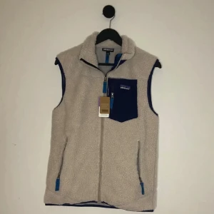 Beige fleeceväst från Patagonia Ny - Säljer en beige fleeceväst från Patagonia med marinblå detaljer. Västen har hög krage, hel dragkedja framtill och tre fickor med blå dragkedjor, varav en bröstficka med logga. Perfekt för lager-på-lager och chill utomhus.