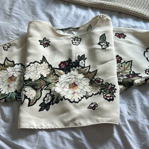 Blommig blus från Zara Basic XS - Supersnygg blus från Zara Basic i ljus beige med stora blommiga print i vitt, grönt och vinrött. Blusen har rund hals och är tillverkad i ett glansigt, mjukt material som ger en lyxig känsla. Perfekt för dig som gillar statement-plagg med coola detaljer.