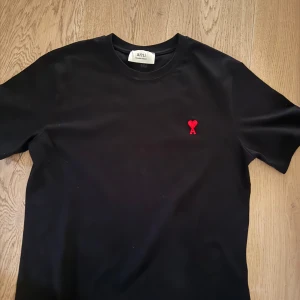 Svart ami t-shirt - Svart t-shirt från Ami med klassisk rund hals och korta ärmar. På bröstet finns ett broderat rött hjärta med bokstaven A. T-shirten är gjord i mjuk bomull och har en clean, stilren look som funkar till jeans eller shorts. Nyttskick och använd fåtal gånger.