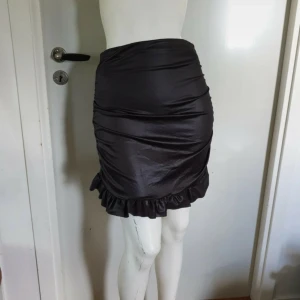 Svart volangkjol från PrettyLittleThing - Snygg svart kortkjol från PrettyLittleThing med glansig finish och volangdetalj längst ner. Kjolen har en tight passform och är gjord i ett syntetmaterial som ger en cool look. Perfekt att styla med boots eller klackar för en trendig vibe.