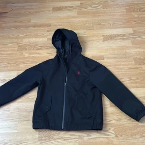 Svart vindjacka från Polo Ralph Lauren - Svart vindjacka med huva från Polo Ralph Lauren. Jackan har en diskret röd logga på bröstet, dragkedja framtill och två fickor med lock. Perfekt för blåsiga dagar och enkel att matcha med olika outfits. Storlek 10-12 år.