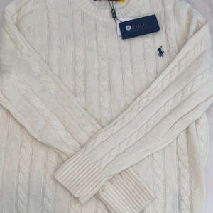 Vit kabelstickad tröja Polo Ralph Lauren - Säljer en vit kabelstickad tröja från Polo Ralph Lauren med klassisk rund hals och broderad marinblå logga på bröstet. Tröjan har långa ärmar och ribbade muddar, perfekt för dig som gillar stilren och tidlös design.