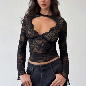 Svart spetsig långärmad topp - Säljer en svart långärmad topp i transparent spets med blommigt mönster. Toppen har en snygg cut-out framtill. Perfekt för dig som vill sticka ut med coola detaljer.