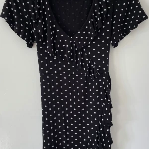 Svart prickig kortklänning  - Volang nertill med. Syns inte på bilden än. Supersöt svart kortklänning från Boohoo med vita prickar, volangdetaljer och knytband i midjan. Klänningen har v-ringning och korta ärmar, vilket ger en chill och lekfull vibe. Perfekt för dig som gillar retrostil och vill sticka ut lite extra.