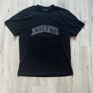 Svart t-shirt Axel Arigato - Svart t-shirt från Axel Arigato med stor vit text 'ARIGATO' broderad över bröstet. Klassisk rund hals och korta ärmar. T-shirten är gjord i mjuk bomull och har en clean, stilren vibe som passar perfekt till jeans eller shorts.