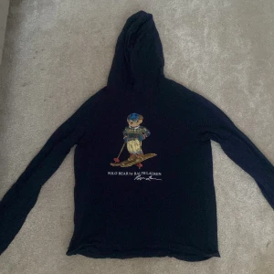 Mörk hoodie från Ralph Lauren med Polo Bear - Säljer en mörk blå hoodie från Ralph Lauren med det ikoniska Polo Bear-motivet på framsidan. Tröjan har långa ärmar och en bekväm huva med dragsko. Perfekt för en avslappnad stil med en touch av lyx. Pris kan diskuteras och kan byta