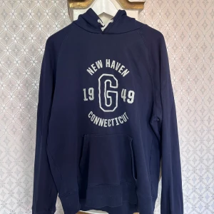 Mörkblå hoodie från GANT med tryck - Snygg mörkblå hoodie från GANT med vit tryckt text 'New Haven Connecticut 1949 G' på bröstet. Tröjan har huva med snörning och en stor magficka. Materialet är mjuk bomull blandat med polyester, perfekt för chill dagar. Klassisk och sportig college-vibe.