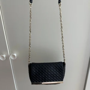 Svart axelväska från Diane von Furstenberg - Snygg svart axelväska i skinn från Diane von Furstenberg med guldfärgad kedja och metall-detalj framtill. Väskan har ett präglat geometriskt mönster på framsidan och en stilren, kompakt form som passar perfekt för att bära det viktigaste