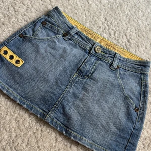 Jeanskjol med gula detaljer Mini Blue - Cool kort jeanskjol från Mini Blue med snygga gula detaljer och nitar på fickorna. Kjolen har klassiska jeansfickor fram och bak, gul insida i midjan och dekorativ knapp framtill. Perfekt för dig som gillar street och denim.