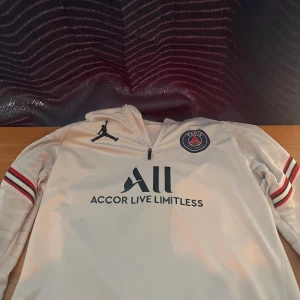 PSG x Jordan vit half zip tröja - Säljer en vit PSG x Jordan tröja med half zip och lång ärm. Tröjan har Paris Saint-Germain-logga på bröstet, Jordan-logga och snygga svarta tryck med Accor Live Limitless och Visit Rwanda. Ränder i rött och blått på ärmarna. Perfekt för fotbollsfans!