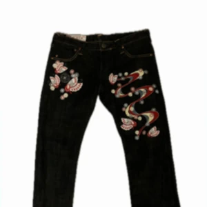 Mörkblåa Selvedge-jeans med broderade fjärilar - Unika mörkblåa jeans från Evisu med färgglada broderade fjärilar och blommor i rött, vitt, blått och gult. Snygga detaljer längs benen och framfickan, klassisk femficksmodell och kontrastsömmar. Perfekt för dig som vill sticka ut med coola mönster och streetstyle. Tveka inte på att höra av er vid frågor!