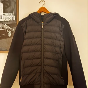 Svart cardigan från 19v69 italia by versace - Svart pufferjacka från Versace 19.69 Italia med huva. Jackan har dragkedja, två fickor med zip och en stilren badge på ärmen. Perfekt för dig som gillar en clean look med italiensk touch. Nypris: 2999